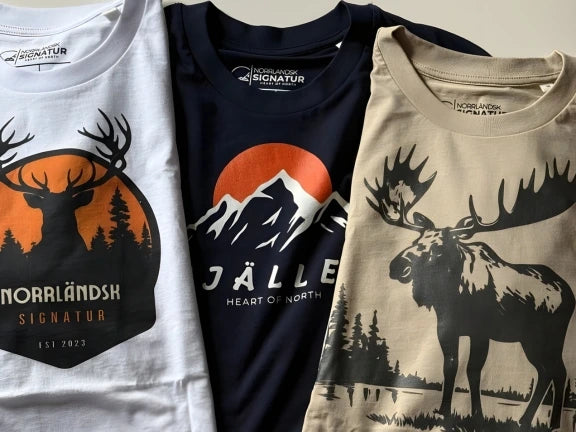 Tre Norrländska streetwear T-shirts från Heart of North Tre Norrländska streetwear T-shirts från Heart of North: en vit med hjortmotiv och texten 'Norrländsk Signatur', en marinblå med fjällmotiv och texten 'JÄLL', samt en beige med älgmotiv – inspirerade av naturen i Norrland.