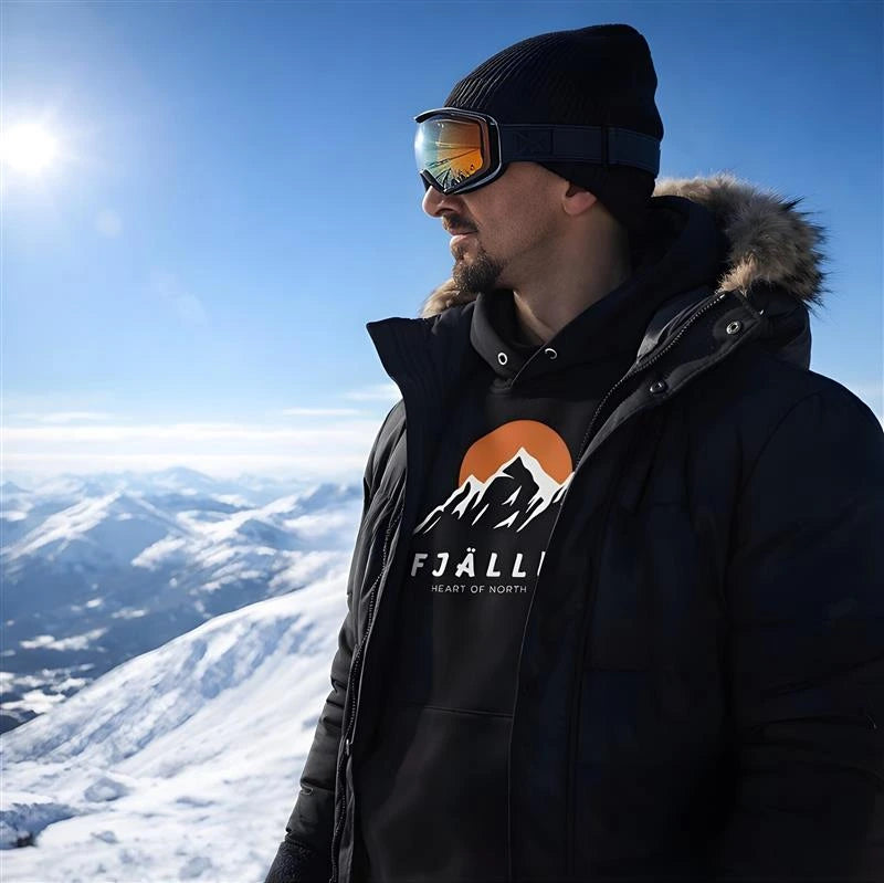 Man i svart hoodie med fjällmotiv från Heart of North står på snötäckt fjälltopp med utsikt över norrländska berg