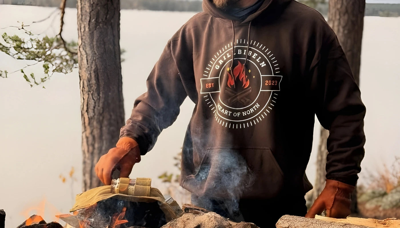 Man grillar vid en öppen eld i norrländsk skog med utsikt över en sjö. Han bär en brun ”Grillbibeln – Heart of North”-hoodie och en svart keps. Veden, elden och naturen runt omkring skapar en äkta norrländsk outdoor-känsla.