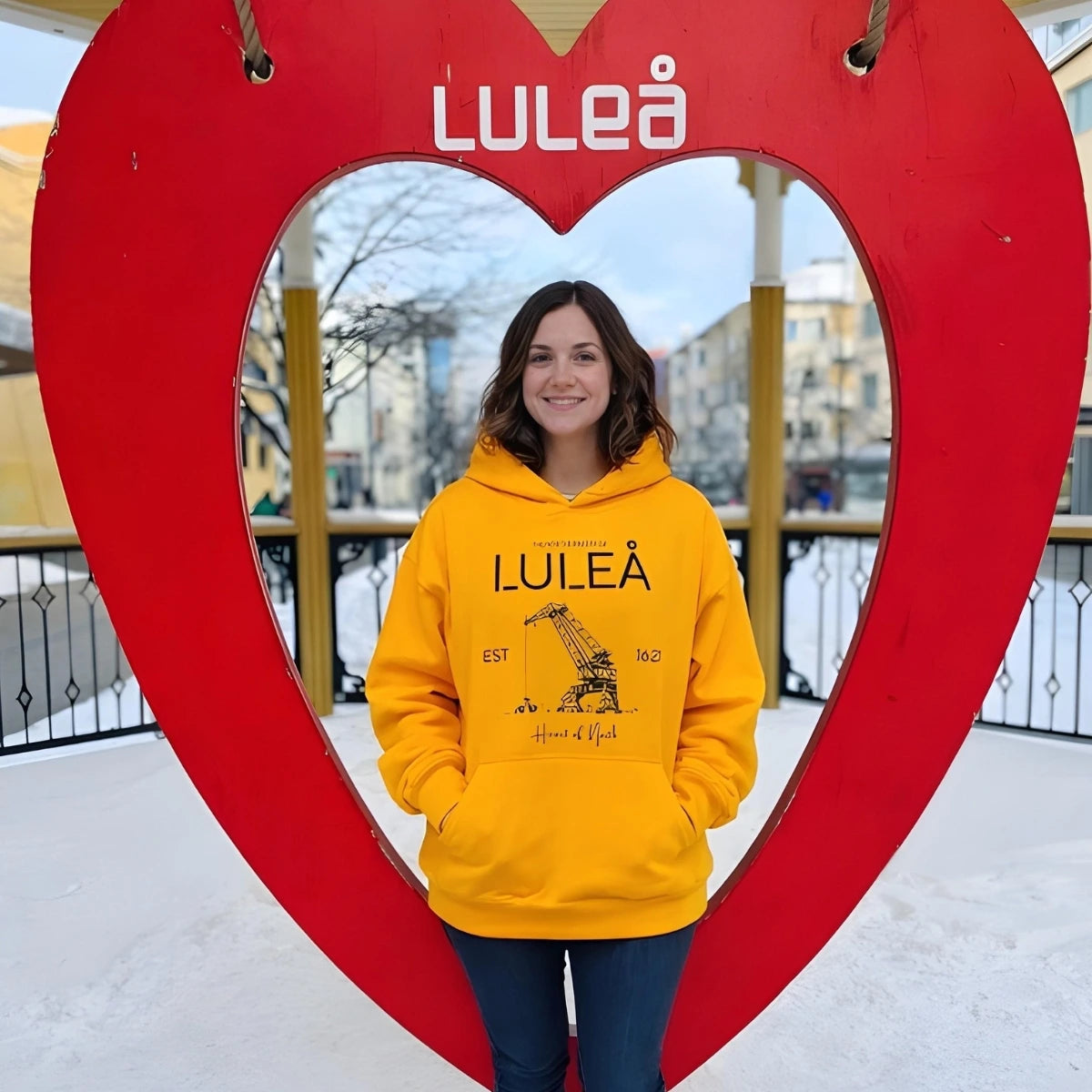 Kvinna i gul hoodie med Luleå motiv från Heart of North står i hjärtformad installation i Luleå vintertid