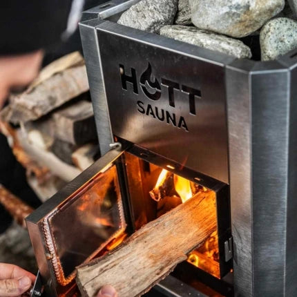 Närbild på vedeldad bastukamin från Hott Sauna med öppen lucka och ved som matas in, omgiven av bastusten och värmande eld.