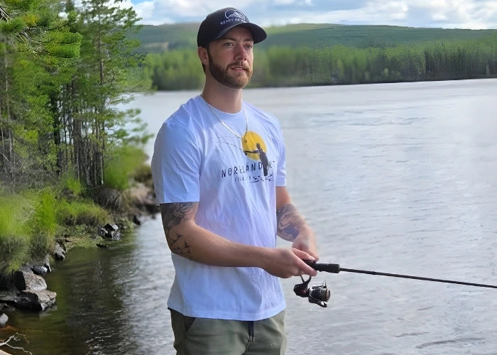 Man står och fiskar vid en norrländsk älv iförd vit t-shirt med fiskemotiv och svart keps från Heart of North – en unisex streetwear-look inspirerad av Norrlands natur.