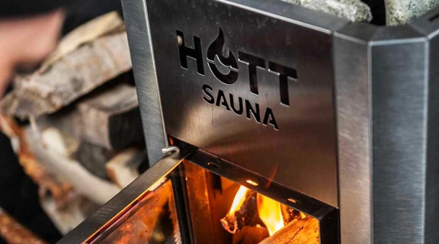 Närbild på vedeldad bastukamin från Hott Sauna med öppen lucka och ved som matas in, omgiven av bastusten och värmande eld.