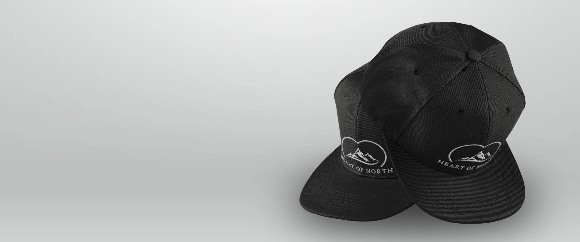 Två svarta kepsar från Heart of North med broderad vit logotyp som föreställer fjäll och texten 'Heart of North' – stilren Norrländsk streetwear-accessoar.