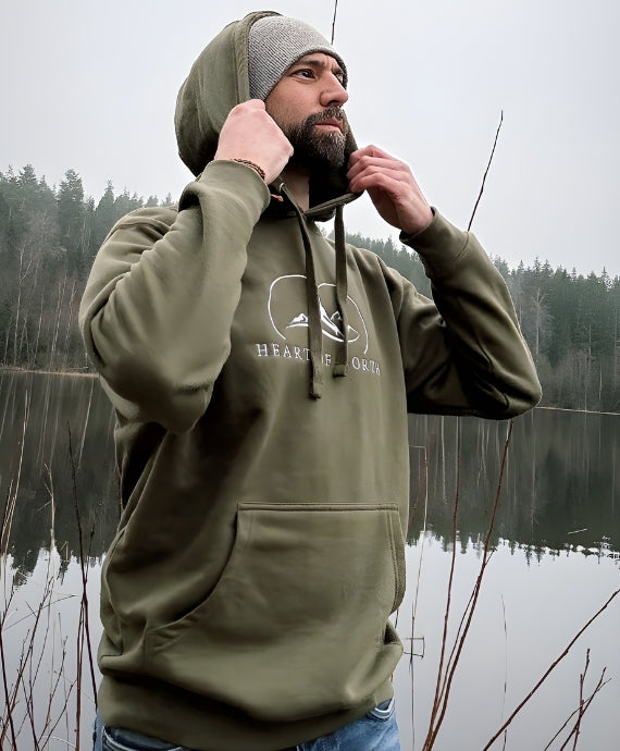 Man i militärgrön hoodie från Heart of North med fjällmotiv och text. Står vid en stilla sjö i norrländsk natur, redo för äventyr i streetwear med utomhuskänsla.