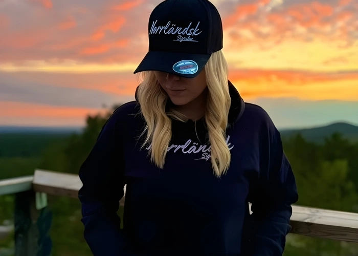 Kvinna i svart hoodie och keps med texten 'Norrländsk' från Heart of North, poserar i solnedgång över fjällandskap