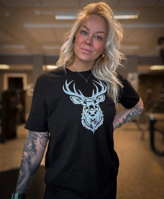 Kvinna i svart T-shirt med vitt hjorthuvud-tryck från Heart of North. Bär Norrländsk streetwear med stolthet i inomhusmiljö, symboliserar styrka och nordlig karaktär.